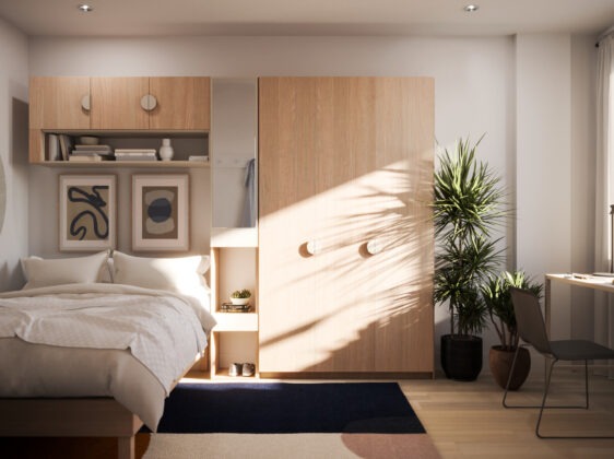 Student bedroom with double bed, beech colored wood shelving and closet, desk and chair under window with sunlight streaming through.